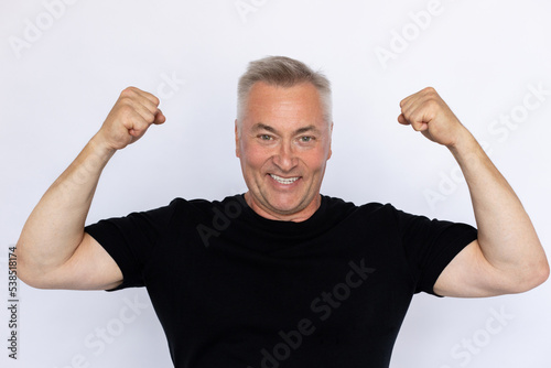 Portrait of happy elderly man showing biceps. Excited Caucasian male model in black T-shirt with gray hair looking at camera, smiling and raising arms. Strength, success concept.