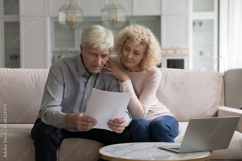 Concerned older couple, wife and husband sit on sofa at home read received formal notification from bank about mortgage denial, unpaid taxes, financial issues, learn insurance terms looking serious