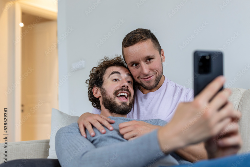 Obraz premium Cute young gay couple video calling their friends in their living room at home. Two male lovers smiling cheerfully while greeting their friends on a smartphone. Young gay couple sitting together.