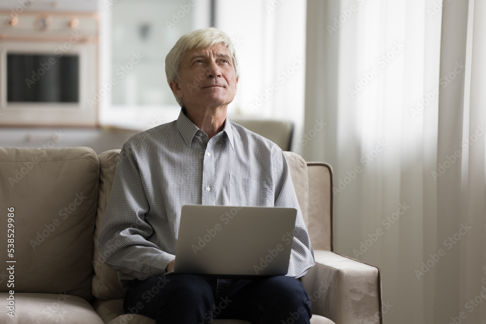 Pensive hoary senior man sits on sofa with laptop, ponder over new blog text, writes letter to friend, e-mailing using modern device and internet connection. Older generation and tech, search ideas