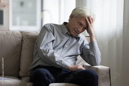 Angry mature hoary man sitting on sofa looking pensive deep in unpleasant thoughts, feels annoyed or dissatisfied by life troubles. Psychological problems, mental disorder, chronic senile diseases