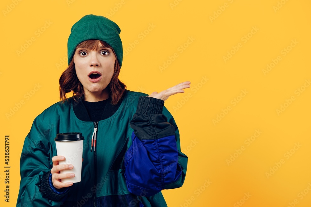 an enthusiastic, surprised woman stands in a green jacket and hat on a yellow background holding a glass of coffee in her hand and spreads her hand to the side with her mouth wide open an enthusiastic, surprised woman stands in a green jacket and hat on a yellow background holding a glass of coffee in her hand and spreads her hand to the side with her mouth wide open