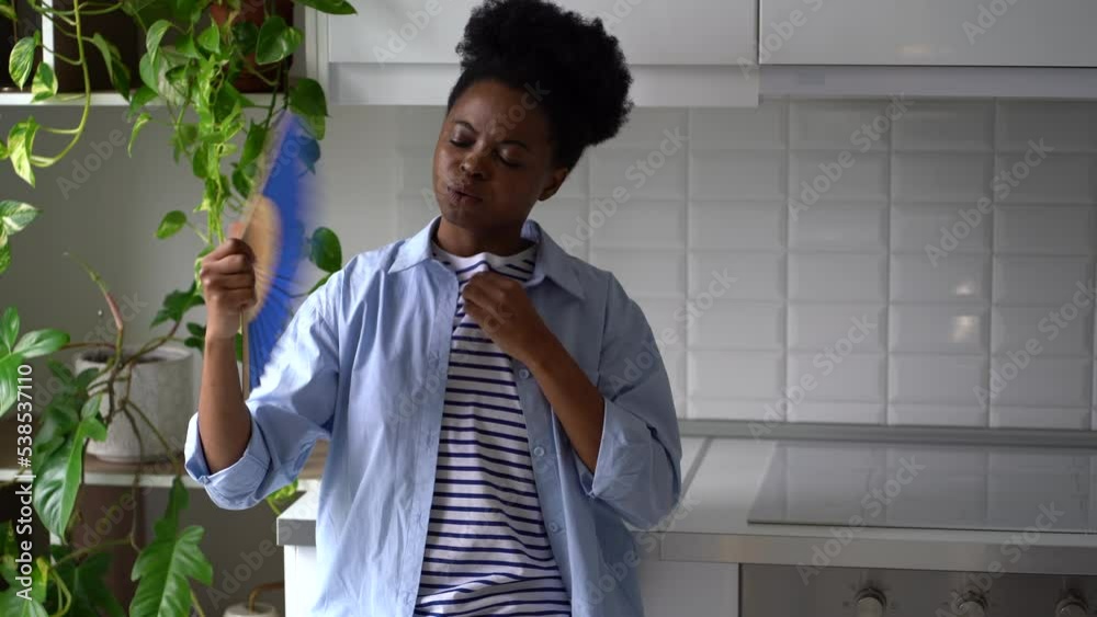 Overheated African American woman trying to cool down with hand fan suffering from lack of air conditioner standing in house. Young black girl feels lethargic due to strong warming and climate change