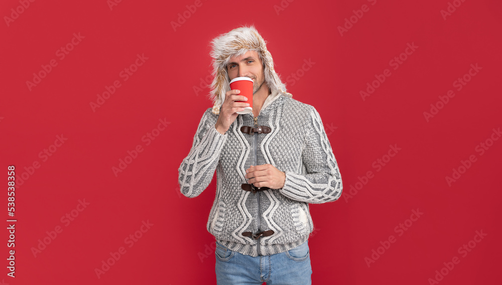 smiling man in sweater and earflap drinking coffee from cup on red background, winter smiling man in sweater and earflap drinking coffee from cup on red background, winter