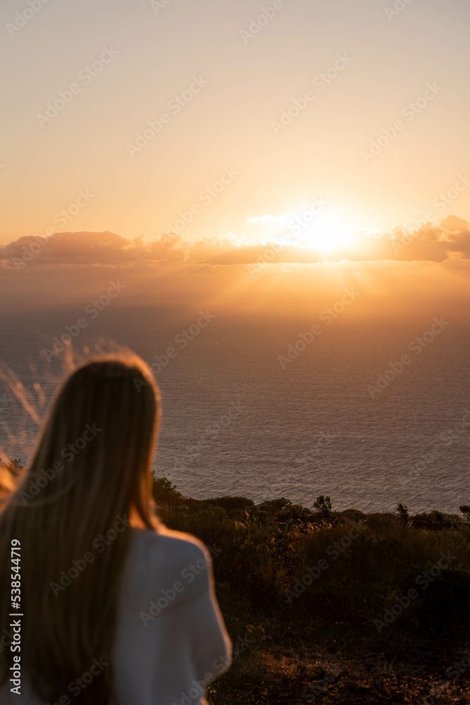 Balckwards of blonde woman in a sunset sky and sun through the clouds.Tranquil seascape and horizon over the water