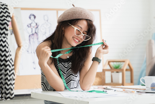 Happy young Asian woman fashion designer or dressmaker wear hat and sunglasses, holding tape measure, smiling and having fun with new idea while working, tailoring tools on table at workplace