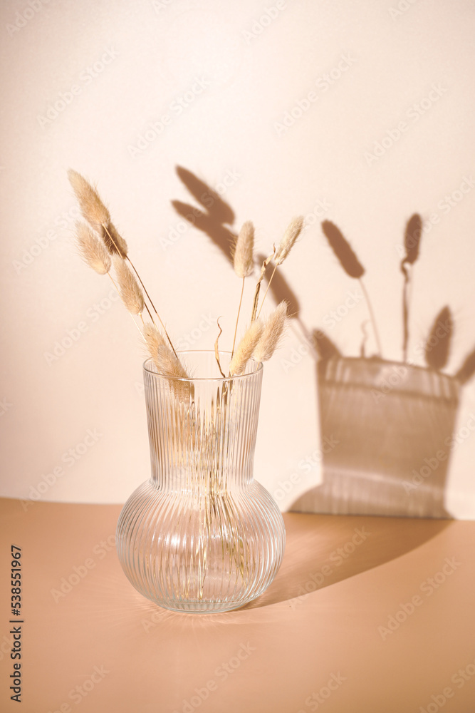 A bunch of yellow wheat spikes, Reeds foliage branches in a transparent rippled vase on orange and beige colored background with hard light shadow, sunny morning
