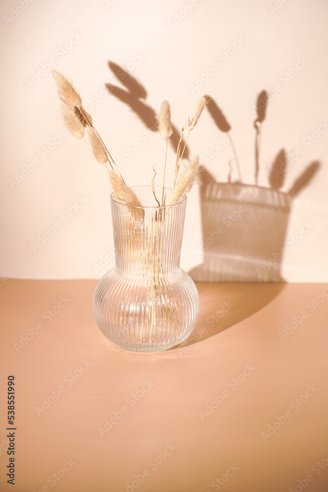 A bunch of yellow wheat spikes, Reeds foliage branches in a transparent rippled vase on orange and beige colored background with hard light shadow, sunny morning A bunch of yellow wheat spikes, Reeds foliage branches in a transparent rippled vase on orange and beige colored background with hard light shadow, sunny morning
