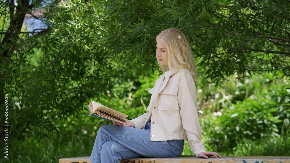 Naklejka premium Carefree young woman reading a fascinating novel while sitting on a park bench.