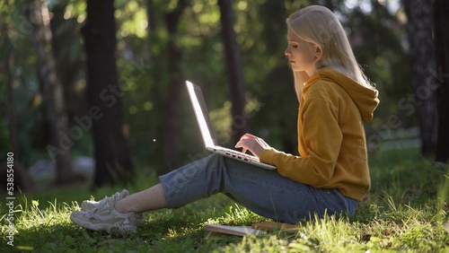 A young blonde woman in casual clothes with a laptop sits on the green grass. The sun illuminates the laptop. Books are lying nearby.