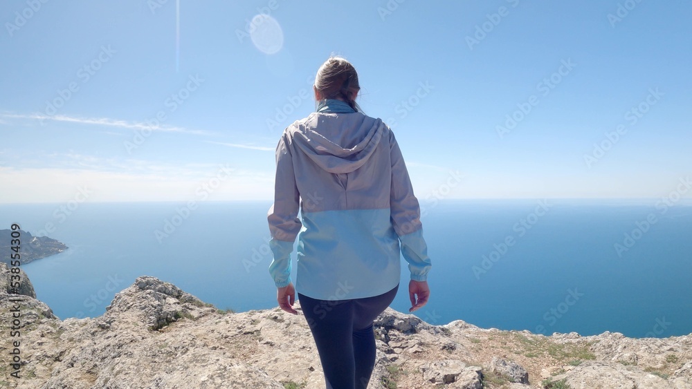 A young woman traveler climbed the highest cliff overlooking the endless ocean. She spread her arms to the sides and enjoys the beautiful view.