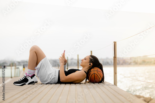 General shot of a basketball player resting, using her smart phone and headphones. with her head on a ball in the harbor photo