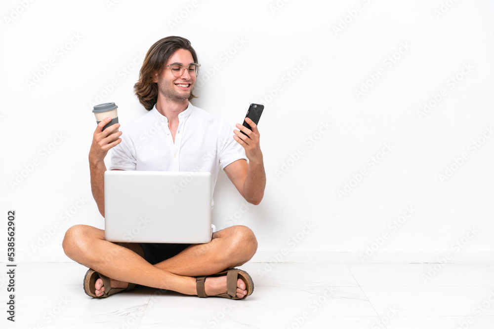 Young handsome man with a laptop sitting on the floor isolated on white background holding coffee to take away and a mobile