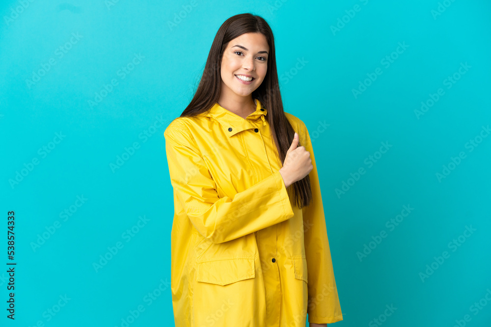 Teenager Brazilian girl wearing a rainproof coat over isolated blue background giving a thumbs up gesture