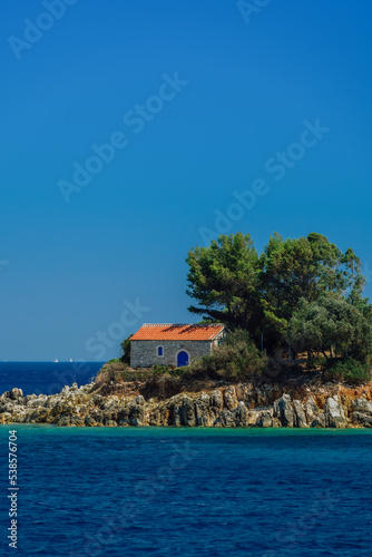 Day view of stone-built Greek Orthodox church with red tiles roof built on a small rocky island.