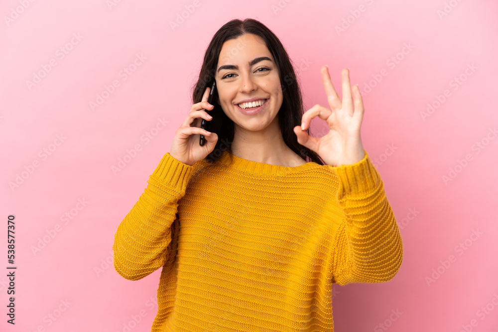 Young caucasian woman using mobile phone isolated on pink background showing ok sign with fingers Young caucasian woman using mobile phone isolated on pink background showing ok sign with fingers