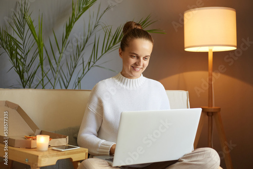 Indoor shot of smiling beautiful woman wearing white sweater sitting in cozy living room and working on laptop, having online job, female freelancer sitting on sofa in front of notebook.