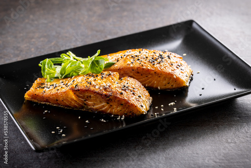 Two salmon fillets baked until crispy, sprinkled with sesame on a black plate. photo