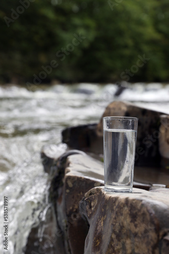 Glass of fresh water on stone near river, space for text