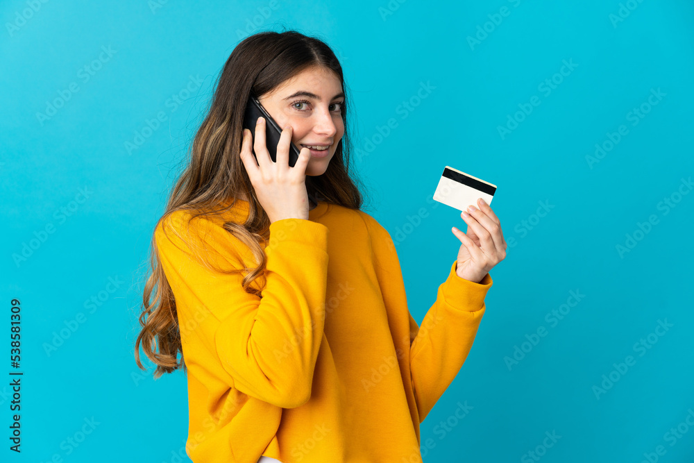 Young caucasian woman isolated on blue background keeping a conversation with the mobile phone and holding a credit card