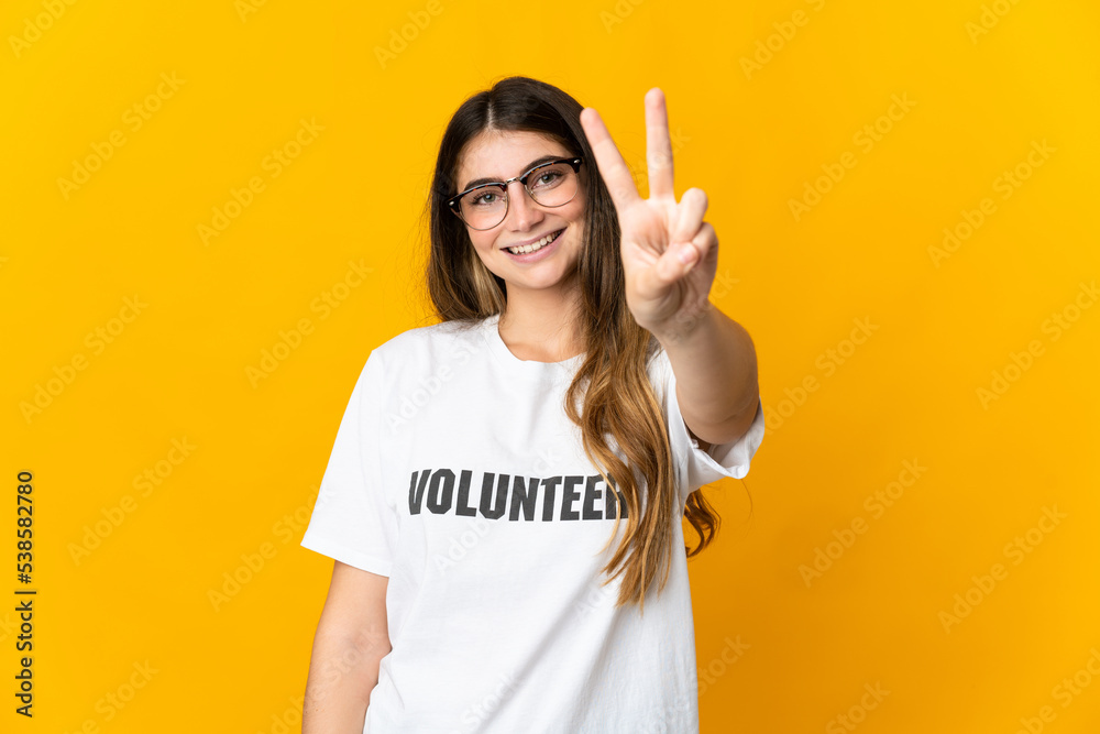 Young volunteer woman isolated on yellow background smiling and showing victory sign Young volunteer woman isolated on yellow background smiling and showing victory sign