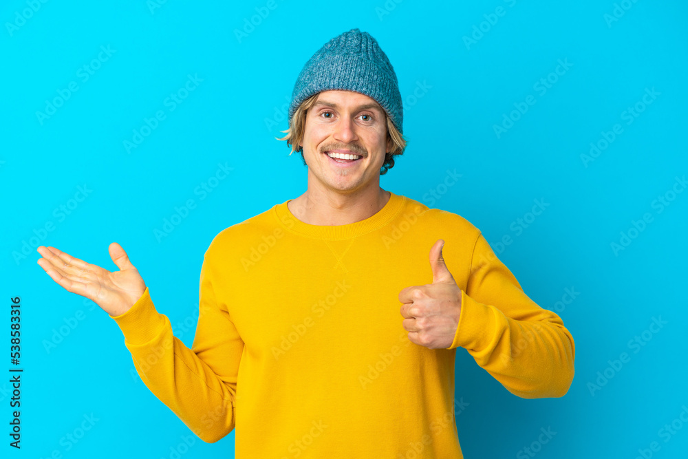 Handsome blonde man isolated on blue background holding copyspace imaginary on the palm to insert an ad and with thumbs up Handsome blonde man isolated on blue background holding copyspace imaginary on the palm to insert an ad and with thumbs up