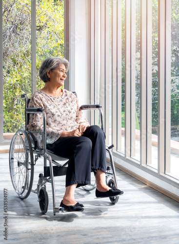 Happy Asian senior woman in casual dress sits in wheelchair, smile and looking out of the glass window with natural green view, vertical style. Positive thinking leads to better health in the elderly.