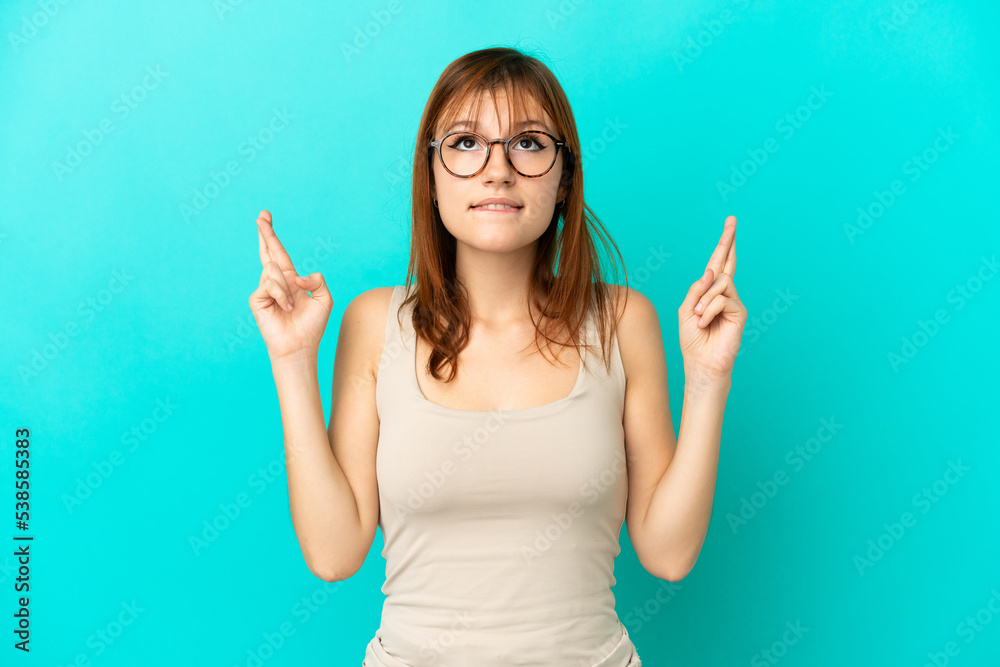 Redhead girl isolated on blue background with fingers crossing and wishing the best Redhead girl isolated on blue background with fingers crossing and wishing the best