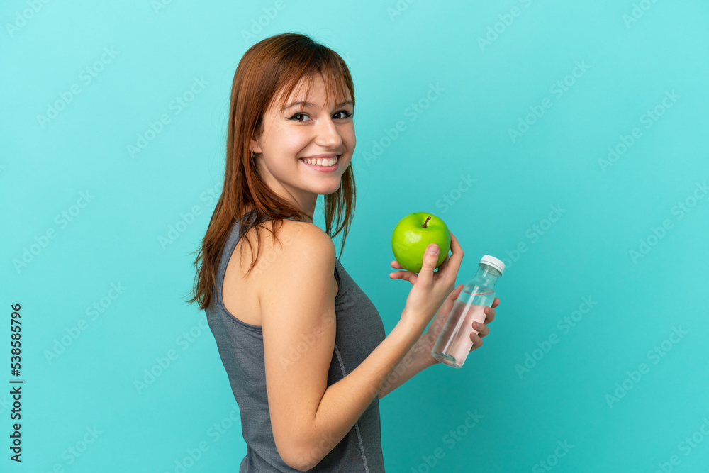 Redhead girl isolated on blue background with an apple and with a bottle of water
