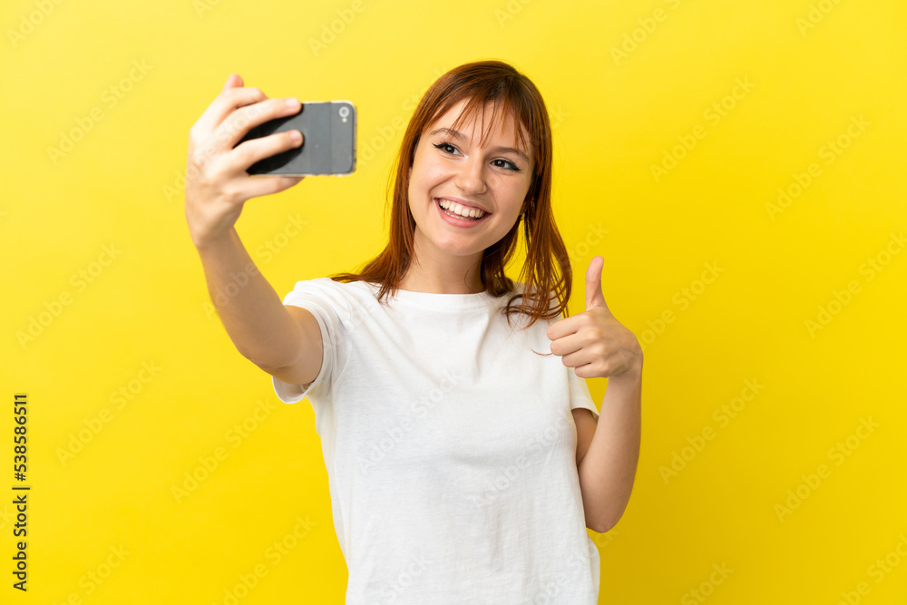 Redhead girl isolated on yellow background making a selfie with mobile phone Redhead girl isolated on yellow background making a selfie with mobile phone