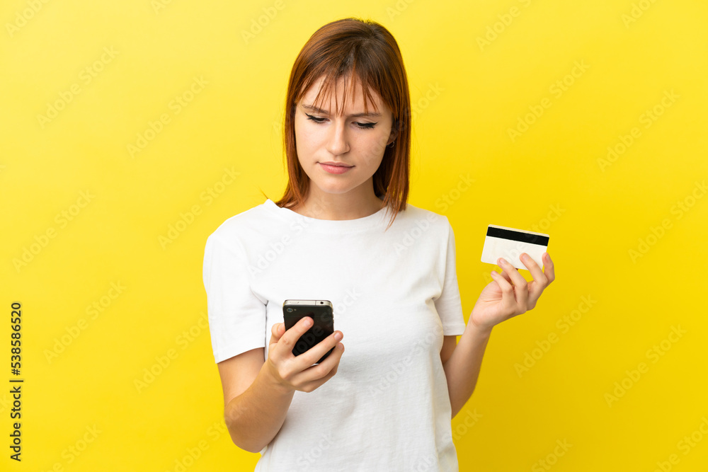 Redhead girl isolated on yellow background buying with the mobile with a credit card Redhead girl isolated on yellow background buying with the mobile with a credit card