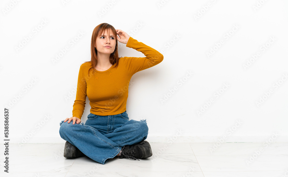 Redhead girl sitting on the floor isolated on white background having doubts and with confuse face expression