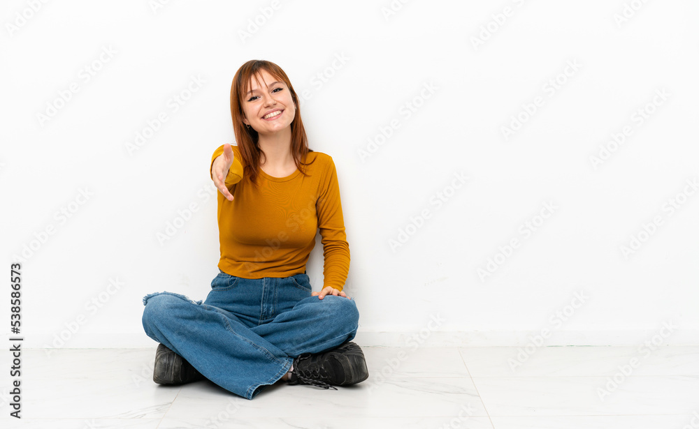 Redhead girl sitting on the floor isolated on white background shaking hands for closing a good deal Redhead girl sitting on the floor isolated on white background shaking hands for closing a good deal