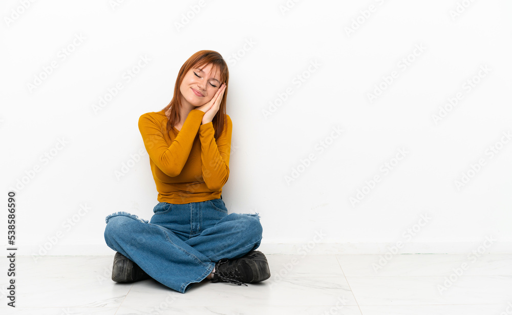 Redhead girl sitting on the floor isolated on white background making sleep gesture in dorable expression Redhead girl sitting on the floor isolated on white background making sleep gesture in dorable expression