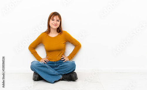 Redhead girl sitting on the floor isolated on white background posing with arms at hip and smiling