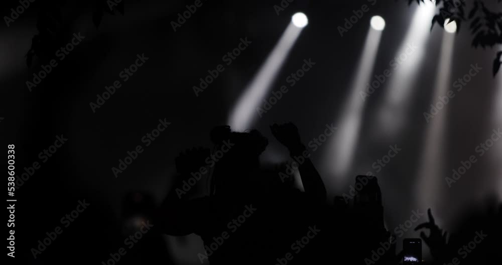 Silhouette people in front of a stage with flashing lights in crowd of people with hands up holding phones and dancing..
