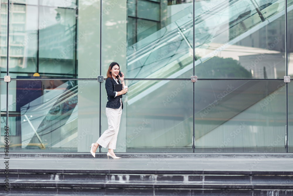 Young Asian businesswoman talking on phone and walking in airport before business trip. Beautiful woman passenger has mobile call and discusses something with smile, holds coffee in hand