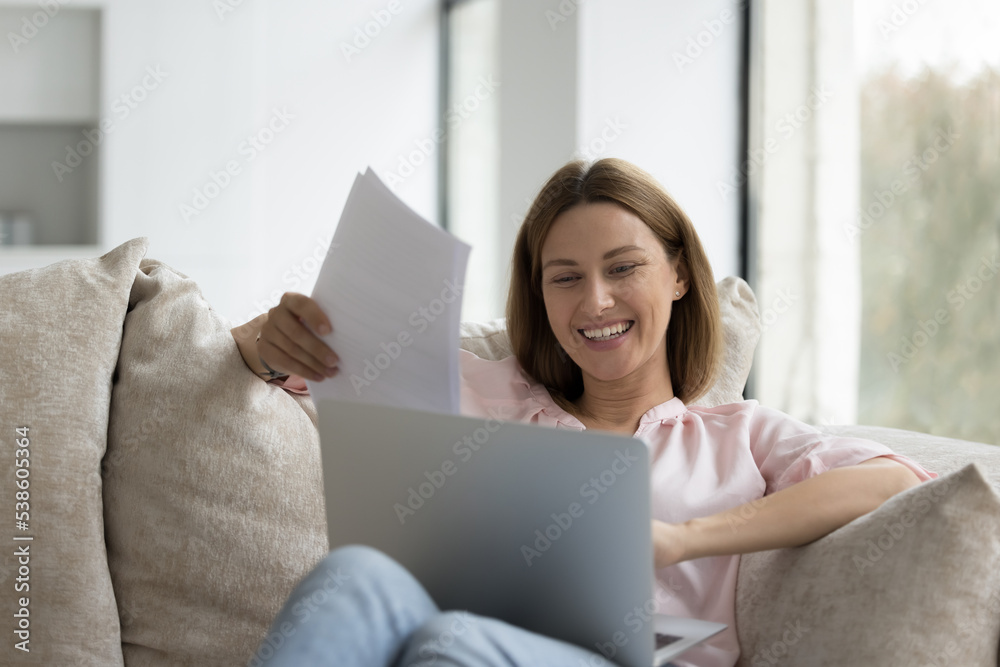 Young 35s woman sits on sofa with laptop, holds bills, makes payment through e-bank application, working at home, doing paperwork smiling looks satisfied, texting on keyboard studying in living room