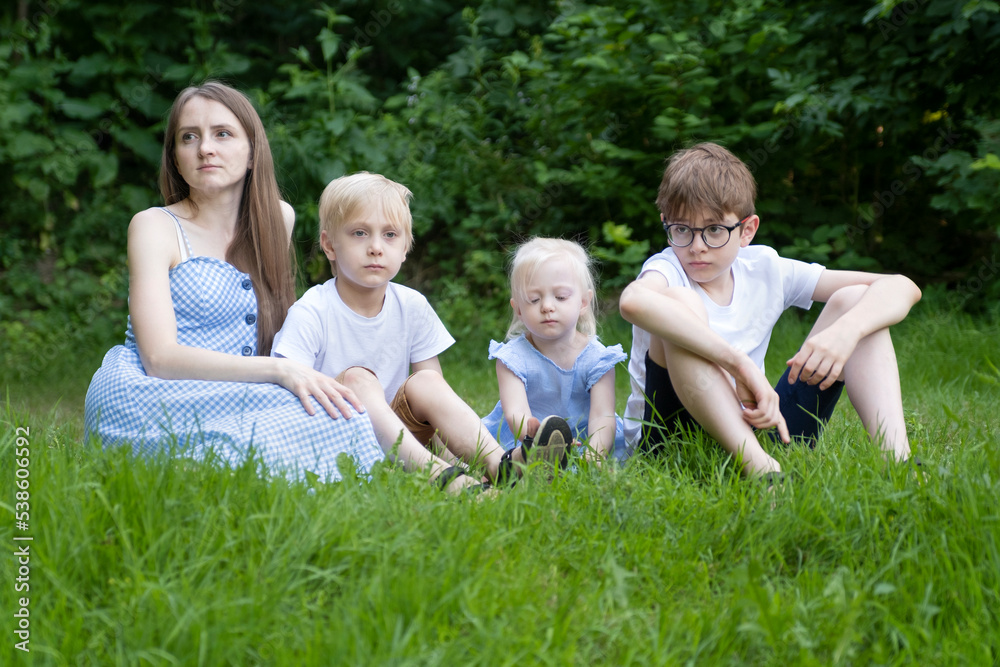 Naklejka premium Woman with three children resting on grass in park. Young mother with daughter and sons sits on green lawn