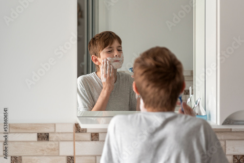 Little brunette boy applies shaving foam to face. Reflection in the mirror. Daily morning procedures