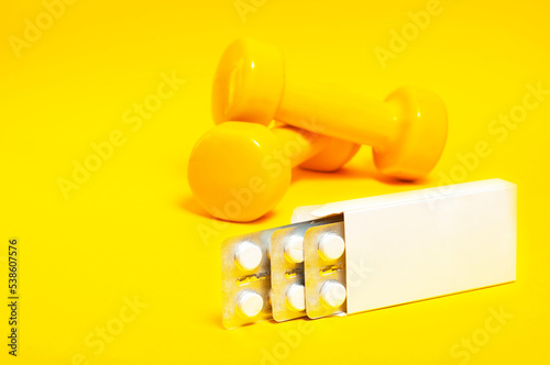 white package of tablets, from which plates with tablets protrude against a background of yellow dumbbells, copy space.