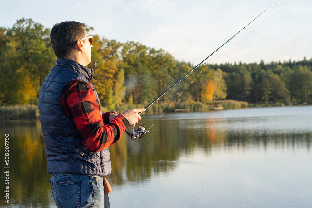 Middle-aged fisherman standing on wooden pier fishing by lake at sunset Middle-aged fisherman standing on wooden pier fishing by lake at sunset