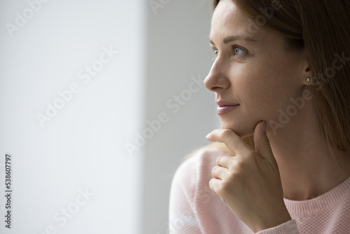 Attractive young 30s woman looks pensive staring into distance, side profile face view with copy space for ad text. Thoughtful female daydreams feels satisfied, ponders, pose alone indoors, close up