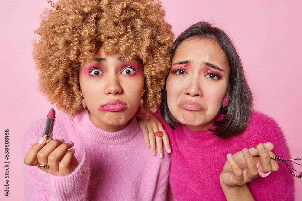 Upset shocked disappointed mixed race women look sadly at camera hold lipstick and eyelashes curler apply makeup undergo beauty procedures isolated over pink background attend makeup courses