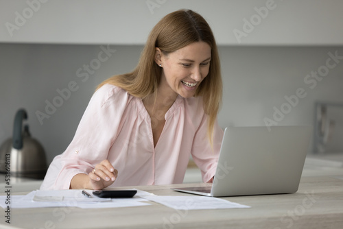 Smiling woman sit at table in kitchen calculates incomes and expenses on calculator, pay utility bills on-line use e-bank on laptop manage family budget, control personal finances feels happy. Savings