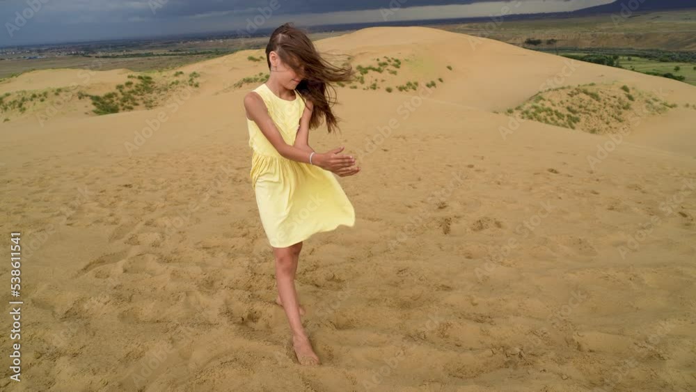 Happy girl in yellow dress stands with sand falling through her hands at sandy dune in desert with mountains and rain clouds on background