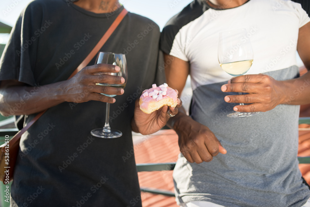 Obraz premium Close-up of African gay mens hands with glasses of champagne. Two men in T-shirts standing close both holding glasses of champagne and one man with cake in his hand. Concept of right for same sex love