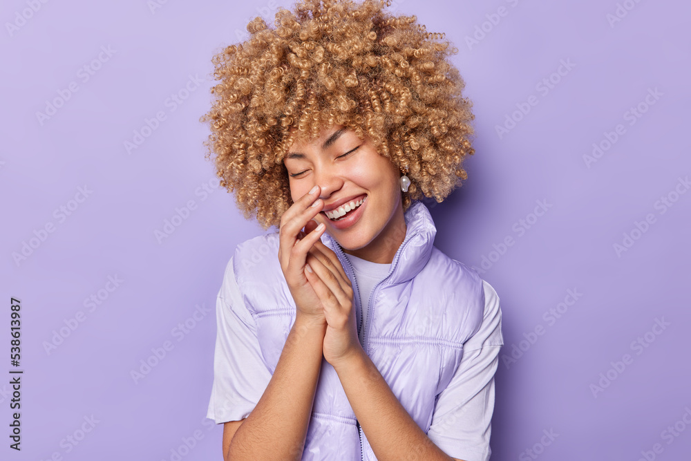 Overjoyed curly haired European woman touches nose smiles happily wears jumper and vest keeps eyes closed expresses positive emotions isolated over purple background. Happiness and joy conept Overjoyed curly haired European woman touches nose smiles happily wears jumper and vest keeps eyes closed expresses positive emotions isolated over purple background. Happiness and joy conept