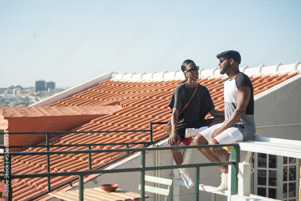 Portrait of Afro American gay men communicating. Two happy boyfriends dating on roof top sitting together on bench talking on blue sky and summer city background. LGBT couples love, relations concept Portrait of Afro American gay men communicating. Two happy boyfriends dating on roof top sitting together on bench talking on blue sky and summer city background. LGBT couples love, relations concept