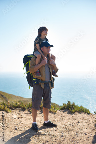 Portrait of happy backpacker standing with son piggyback outdoors. Man and boy hiking in summer. Active family weekend concept